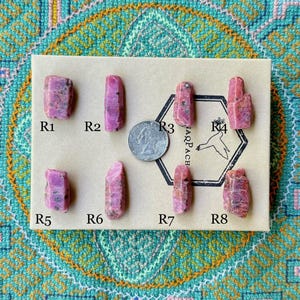 May include: Eight small pink ruby crystals labelled R1 to R8 on a brown card with the text "IAQPACH" and a US quarter.