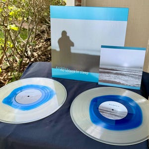 May include: A collection of vinyl records and album art. Two clear vinyl records with blue and white designs are in the foreground. The album art features a silhouette on a beach and the text "Aartíka no solace in sleep."