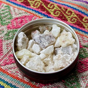 May include: A small, round tin with a silver rim, filled with irregular, off-white and light brown resin pieces. The tin rests on a colourful textile with pink, green, and orange patterns.