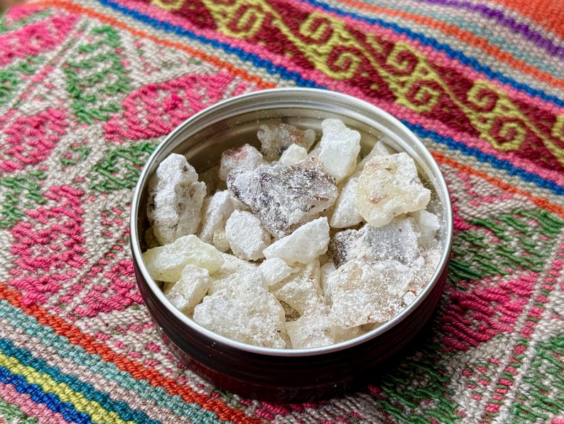 May include: A small, round tin with a silver rim, filled with irregular, off-white and light brown resin pieces. The tin rests on a colourful textile with pink, green, and orange patterns.