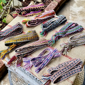 May include: A collection of colorful woven belts with intricate patterns and fringed ends. The belts feature a variety of colors, including purple, red, blue, and yellow, and are displayed on a tan fabric.