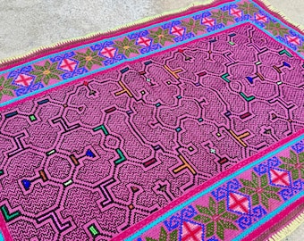 May include: A rectangular, hand-woven textile with a vibrant pink central design featuring geometric patterns. The border is a combination of pink, turquoise, and green, with repeating floral and geometric motifs. The edges have a fringe.