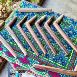 Könnte beinhalten: Eine Sammlung handgefertigter Haarstäbe aus Holz in verschiedenen Brauntönen, angeordnet auf einem farbenfrohen, gemusterten Textil. Die Haarstäbe haben ein gebogenes, L-förmiges Design, das die natürliche Holzmaserung hervorhebt. Der Stoff zeigt eine lebendige Mischung aus Grün, Blau, Rosa und Gelb.