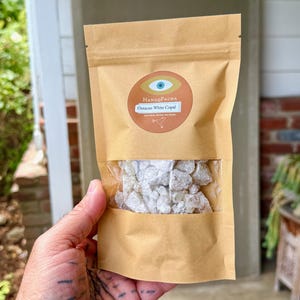 May include: A brown paper bag with a clear window, filled with white copal resin. The bag has a circular label with the text "HanaqPacha" and "Oaxacan White Copal". The background includes greenery and a brick wall.
