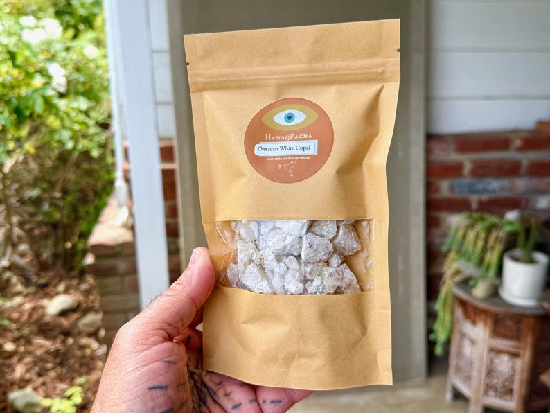 May include: A brown paper bag with a clear window, filled with white copal resin. The bag has a circular label with the text "HanaqPacha" and "Oaxacan White Copal". The background includes greenery and a brick wall.