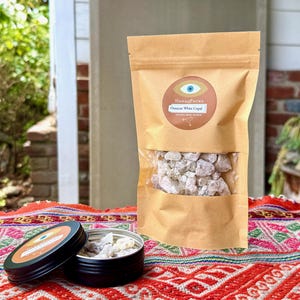 May include: A brown paper bag with a clear window showing white copal resin, and a black tin with a matching label. The label reads "HanaqPacha" and "Oaxacan White Copal". The items are on a colourful patterned textile.