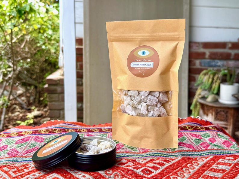 May include: A brown paper bag with a clear window showing white copal resin, and a black tin with a matching label. The label reads "HanaqPacha" and "Oaxacan White Copal". The items are on a colourful patterned textile.