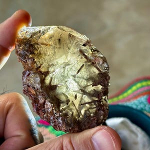 May include: A translucent, golden-yellow gemstone with dark inclusions is held between fingers. The rough-cut stone has a crystalline structure and a reddish-brown outer layer. The background is blurred, with a glimpse of colorful textiles.