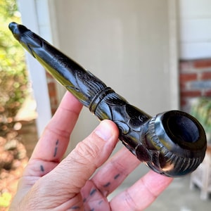May include: A carved black smoking pipe with floral and bird designs. The pipe is held in a hand, highlighting the detailed carvings and the smooth, polished finish. The pipe's bowl is round and deep.