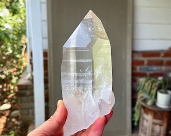May include: A clear quartz crystal point with a faceted, tapered shape. The crystal is held in a hand and has a natural, rough base. The quartz is translucent with subtle internal striations and a slight yellow hue.