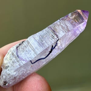 May include: A natural amethyst crystal with a pointed tip and a light purple hue. The crystal has a clear, textured surface with a dark blue line drawn on it. The crystal is held in a person's hand.
