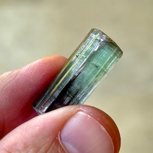 May include: A translucent, elongated gemstone with a mix of green and gold hues. The crystal is held between fingers, showcasing its natural, raw form. The gemstone's surface has visible lines and imperfections, highlighting its unique character.