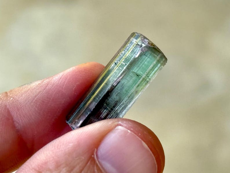 May include: A translucent, elongated gemstone with a mix of green and gold hues. The crystal is held between fingers, showcasing its natural, raw form. The gemstone's surface has visible lines and imperfections, highlighting its unique character.