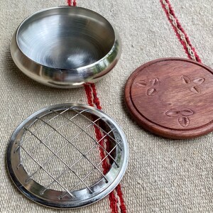 K&ouml;nnte beinhalten: Ein silberner Metall-R&auml;ucherofen mit einem Drahtgitter-Einsatz und einem Holzdeckel mit einem geschnitzten Design. Der Deckel hat drei geschnitzte Kaffeekorn-Designs.