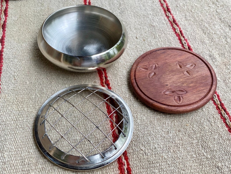 K&ouml;nnte beinhalten: Ein silberner Metall-R&auml;ucherofen mit einem Drahtgitter-Einsatz und einem Holzdeckel mit einem geschnitzten Design. Der Deckel hat drei geschnitzte Kaffeekorn-Designs.