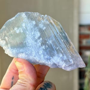 May include: A translucent, light blue selenite crystal formation. The crystal has a layered appearance with visible striations and a slightly rough texture. The overall shape is irregular, with a natural, unpolished look. The crystal is held in a hand.