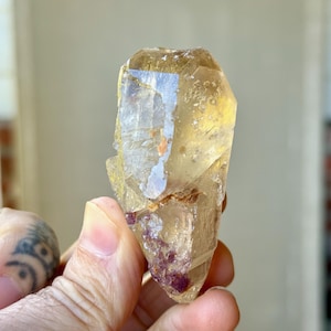 May include: A translucent, yellow-toned crystal with a faceted, elongated shape. The crystal has a clear, glassy appearance with some internal inclusions and a touch of reddish-purple at the base. The crystal is held in a hand.