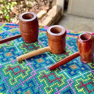 May include: Three wooden pipes with cylindrical bowls and long stems are displayed on a colourful, patterned textile. The pipes are made of polished wood, with variations in colour and grain. The background is blurred, suggesting an outdoor setting.