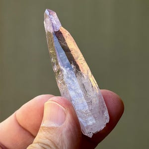 May include: A translucent, elongated quartz crystal point with a pale purple amethyst hue. The crystal features a sharp, pointed termination and a faceted, prismatic structure with visible internal inclusions and natural surface textures.