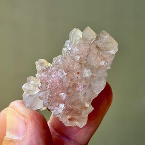 May include: A cluster of translucent, light pink and clear crystals. The crystals have a rough, textured surface and are clustered together in a natural formation. The crystals are held by a person's fingers.