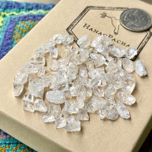Puede incluir: Un conjunto de cristales de cuarzo transparentes y en bruto se apilan sobre una caja beige. La caja tiene un logotipo con el texto "Hana Pacha" y una moneda. Los cristales están dispersos.