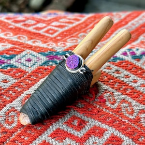 Puede incluir: Un instrumento de madera con dos piezas cilíndricas de madera, envuelto en cuero negro y adornado con una gema púrpura. El instrumento descansa sobre un textil con estampado rojo y naranja.