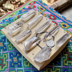 Puede incluir: Nueve puntas de cristal de cuarzo transparente se exhiben sobre una superficie beige, dispuestas en una cuadrícula. Los cristales varían en tamaño y forma, algunos parecen más translúcidos que otros. Se coloca una moneda cerca del centro. Los cristales se presentan sobre un textil colorido y estampado.