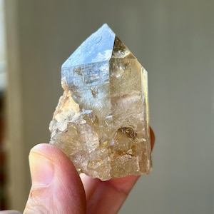 May include: A clear, light brown quartz crystal with a pointed top. The crystal has a rough, textured base. The crystal is a natural mineral specimen, and it is a light brown colour. The crystal is held in a person's hand.