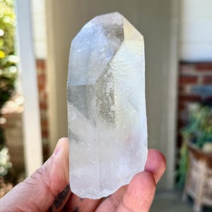 May include: A clear quartz crystal point with a faceted top. The crystal is held in a hand and appears translucent with some internal inclusions. The background is blurred, suggesting an outdoor setting. The crystal is a natural, raw gemstone.
