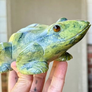 以下が含まれることがあります： 緑と青の色合いの彫刻された石のカエルの彫刻。カエルは、彫刻された脚や茶色の目を持つ詳細な顔など、詳細な特徴を持っています。彫刻は装飾品です。