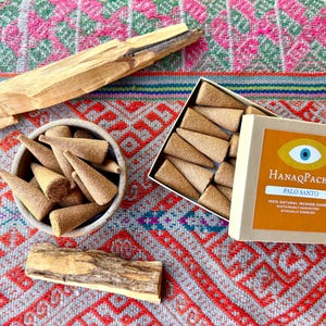 May include: A collection of light brown cone-shaped incense pieces, two pieces of raw palo santo wood, and a square cardboard box labeled HanaqPacha Palo Santo with an eye graphic, containing additional incense cones inside the open container.