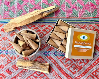 Könnte beinhalten: Eine Sammlung hellbrauner kegelförmiger Räucherkegel, zwei Stücke rohes Palo-Santo-Holz und eine quadratische Pappschachtel mit der Aufschrift HanaqPacha Palo Santo und einem Augensymbol, die weitere Räucherkegel im offenen Behälter enthält.