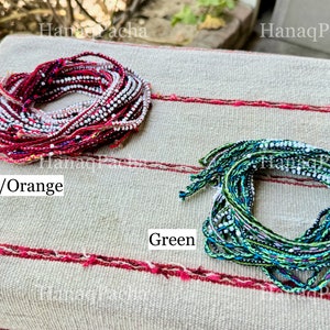 K&ouml;nnte beinhalten: Zwei B&uuml;ndel bunter Fadenarmb&auml;nder, eines rot und orange, das andere gr&uuml;n, mit eingewebten wei&szlig;en Perlen. Die Armb&auml;nder liegen auf einem gestreiften Stoffhintergrund.