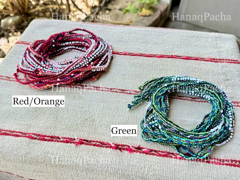K&ouml;nnte beinhalten: Zwei B&uuml;ndel bunter Fadenarmb&auml;nder, eines rot und orange, das andere gr&uuml;n, mit eingewebten wei&szlig;en Perlen. Die Armb&auml;nder liegen auf einem gestreiften Stoffhintergrund.