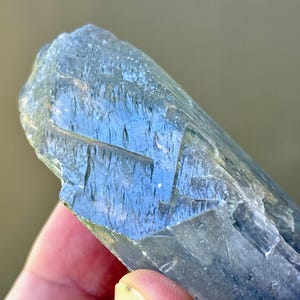 May include: A close-up of a blue-gray crystal with a rough, textured surface. The crystal has a layered appearance with visible striations and a translucent quality, reflecting light. The crystal is held in a hand.