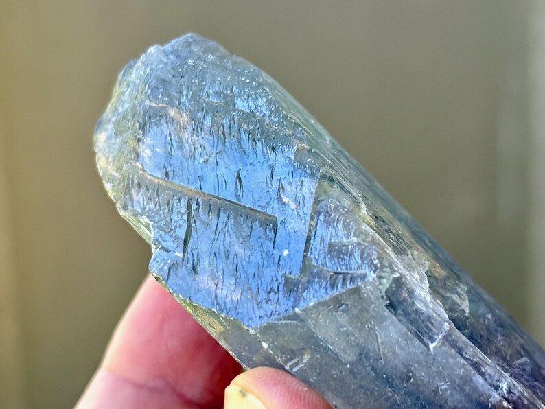 May include: A close-up of a blue-gray crystal with a rough, textured surface. The crystal has a layered appearance with visible striations and a translucent quality, reflecting light. The crystal is held in a hand.