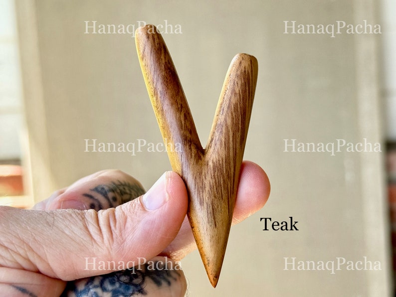 May include: A V-shaped wooden tool with a smooth, polished finish and visible dark brown grain patterns. The object is labeled with the text Teak and features a pointed bottom end with two upward-extending prongs of slightly different lengths.