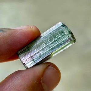 May include: A raw, natural tourmaline crystal. The gemstone is a translucent, rectangular shape with a mix of green, pink, and clear colors. The crystal is held between two fingers, showcasing its natural texture and facets.