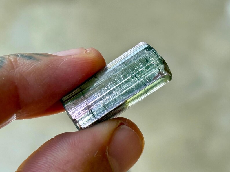 May include: A raw, natural tourmaline crystal. The gemstone is a translucent, rectangular shape with a mix of green, pink, and clear colors. The crystal is held between two fingers, showcasing its natural texture and facets.