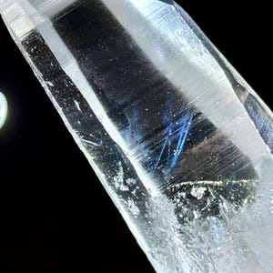 以下が含まれることがあります： 内部に青色のインクルージョンが入った透明な水晶のクローズアップ。結晶は鋭く角度がつけられており、光を反射してキラキラとした効果を生み出しています。明るい円形の光源が、黒い背景に対して背景に見えます。
