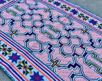 May include: A rectangular textile piece featuring a light pink background with a complex, repeating geometric pattern in dark purple, bright yellow, and neon green. The border displays a white band with colorful floral and star motifs.