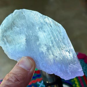 May include: A translucent, light blue crystal formation with a layered, fibrous texture. The crystal is held by a hand, showcasing its natural, irregular shape and subtle color variations. The overall appearance suggests a raw, unpolished gemstone.
