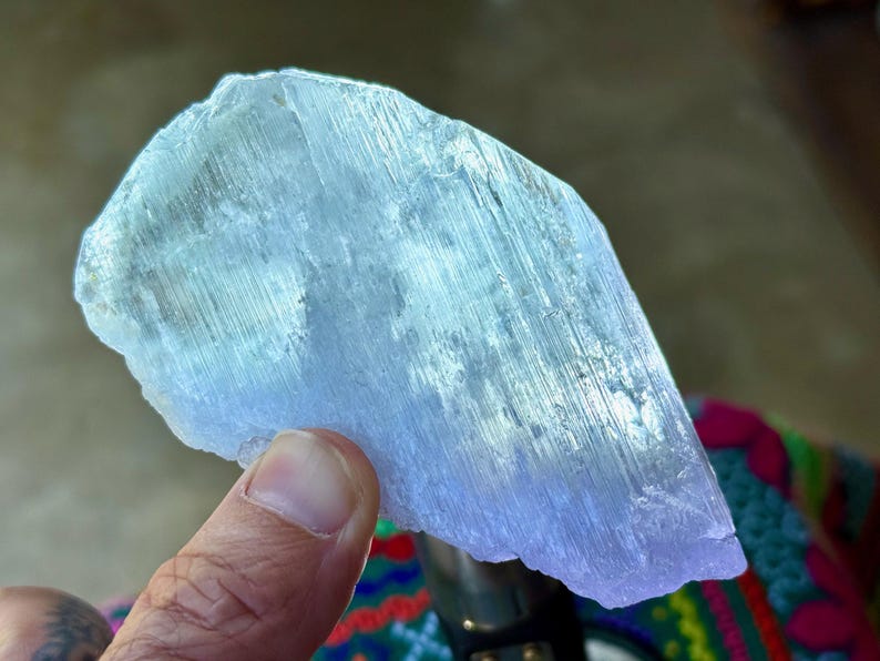 May include: A translucent, light blue crystal formation with a layered, fibrous texture. The crystal is held by a hand, showcasing its natural, irregular shape and subtle color variations. The overall appearance suggests a raw, unpolished gemstone.