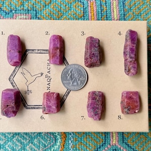 May include: Eight raw ruby crystals in varying shapes and sizes are displayed on a cardboard surface. A US quarter is included for size reference. The crystals range in color from deep pink to reddish-purple, with visible natural textures.