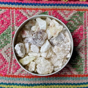 May include: A round, silver tin filled with chunks of off-white and amber-colored resin. The resin pieces vary in size and texture, some appearing crystalline. The tin sits on a colorful, patterned textile with pink, green, and blue hues.