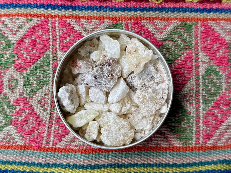 May include: A silver tin containing pieces of off-white and amber resin. The resin chunks are of varying sizes and textures, some appearing crystalline. The tin rests on a colourful textile with pink, green, and blue patterns.