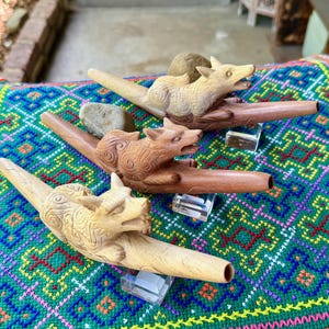 May include: Four carved wooden pipes with animal head designs, including a fox and a wolf. The pipes are displayed on a colorful, patterned textile with green, blue, and pink hues. Each pipe rests on a clear stand.