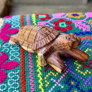 May include: A hand-carved wooden turtle figurine with detailed shell and body features. The turtle is a warm brown color and rests on a colorful, patterned textile with pink, green, yellow, and blue hues. The figurine is approximately 6 inches long.
