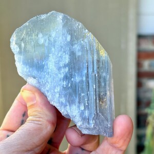 May include: A light blue crystal with a rough, textured surface. The crystal is held in a hand, showcasing its natural form and color variations. The crystal's shape is irregular, with visible striations.