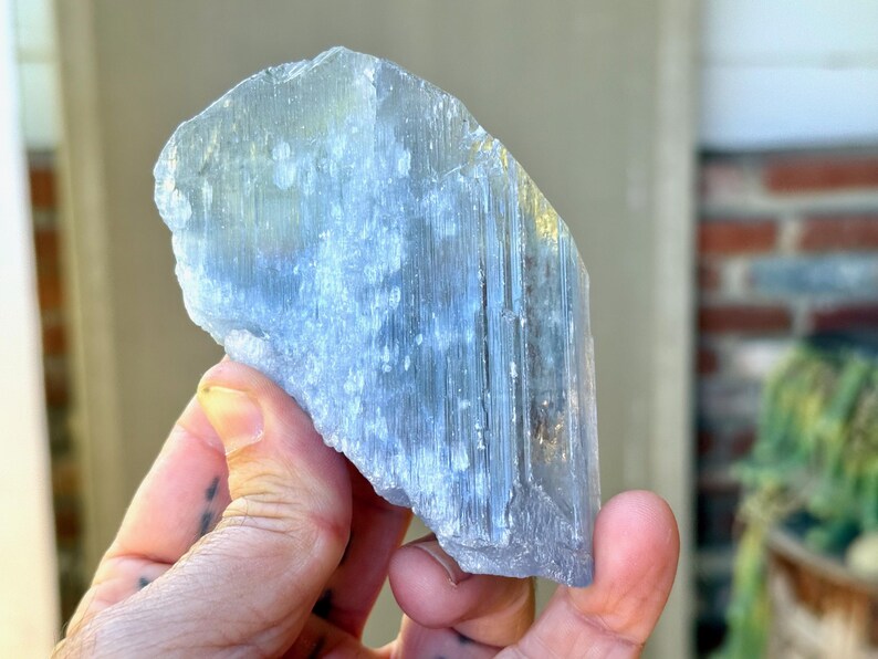 May include: A light blue crystal with a rough, textured surface. The crystal is held in a hand, showcasing its natural form and color variations. The crystal's shape is irregular, with visible striations.